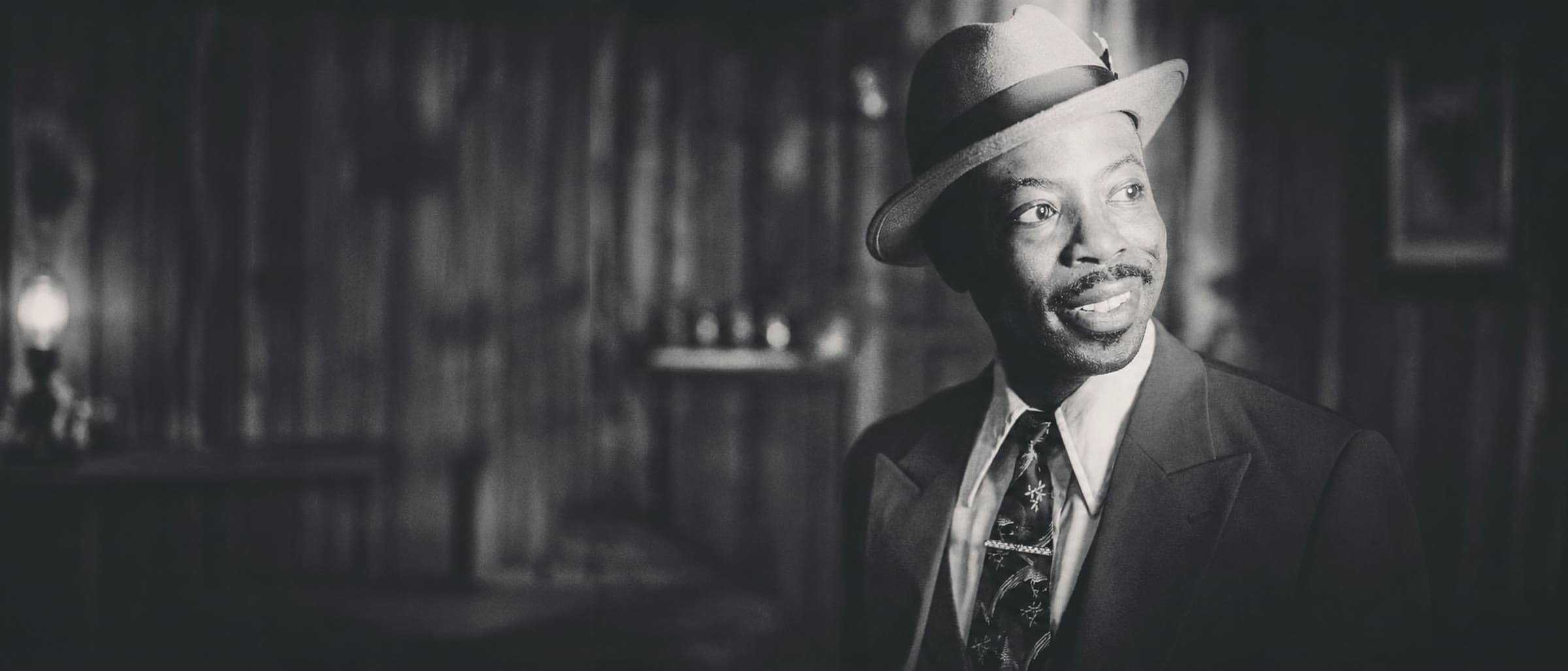 Charlie Hudson III in a period suit and hat, photographed in a dimly lit interior.